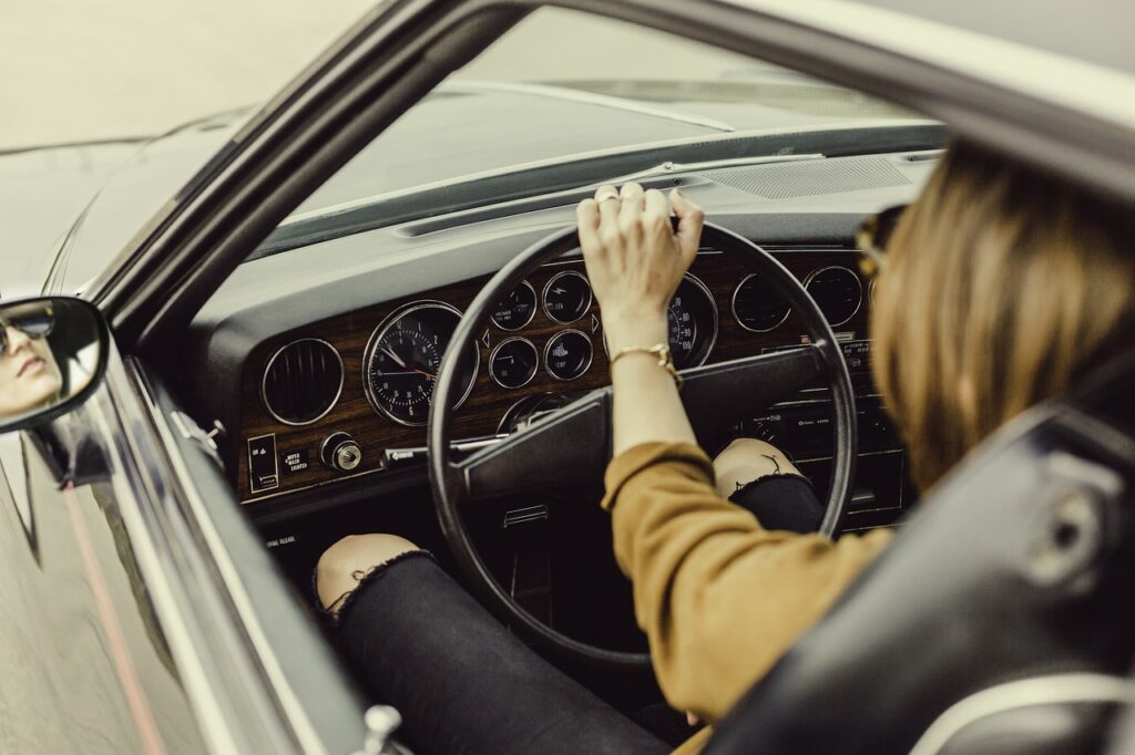 Person driving a car with one hand on the steering wheel and the other on the gear shift, as seen from the passenger seat.