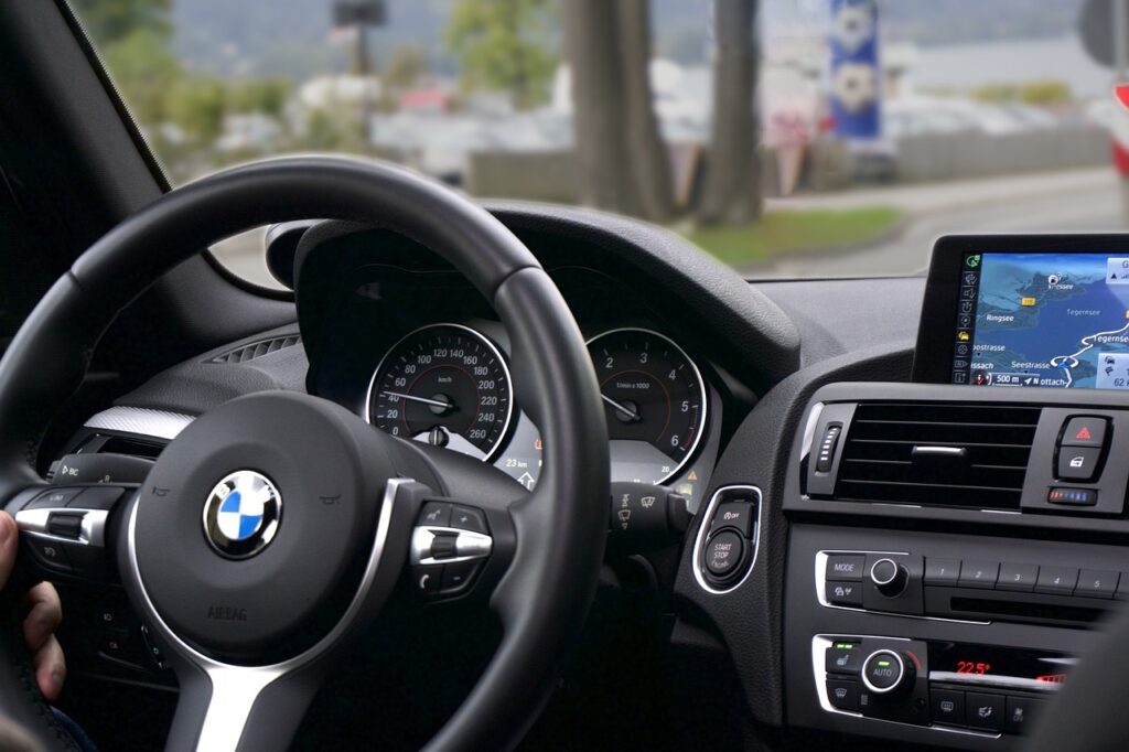 Interior of a BMW car, showing the steering wheel, speedometer, and GPS navigation system on the dashboard screen, with an outside view of trees and a lake.