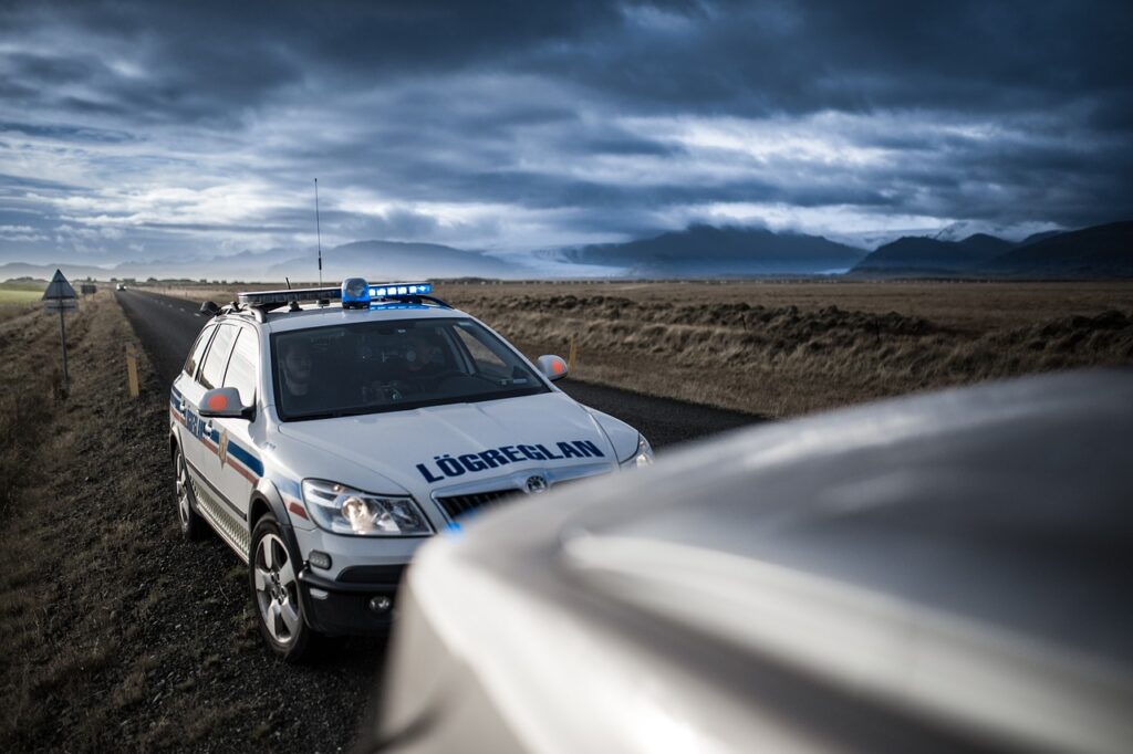 A police car with flashing lights is parked on the side of a rural road, with mountains and a cloudy sky in the background.