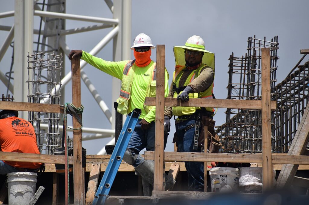 Two construction workers in safety gear stand at a construction site, one leaning on a wooden frame and the other holding a hammer. Rebar structures and a ladder are visible.