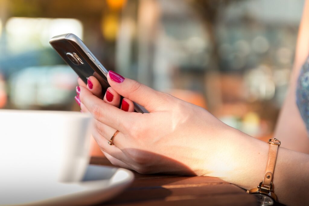 A person with painted nails holds a smartphone in one hand and rests the other on a table, with a coffee cup in the foreground.