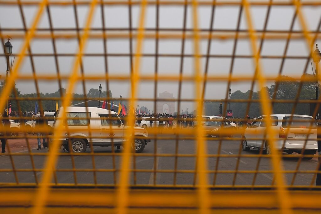 View through a yellow barricade showing cars and people gathered on a road with a prominent monument visible in the background.