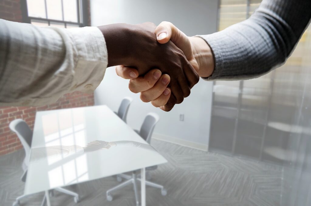 Two individuals shaking hands over a desk in a modern office setting with chairs and a window in the background.