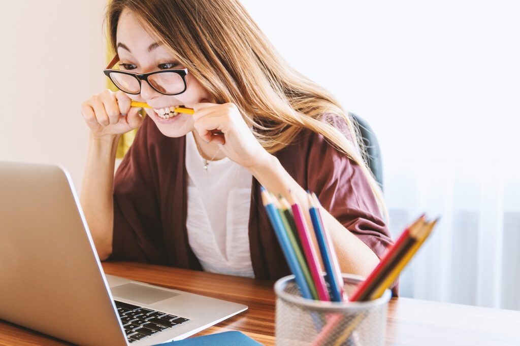 A woman wearing glasses is sitting at a desk, looking at a laptop screen while biting a pencil. Colored pencils are in a container nearby.