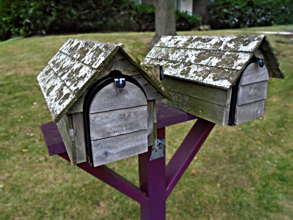Two weathered wooden mailboxes with mossy roofs on a single purple post, set in a grassy outdoor area.