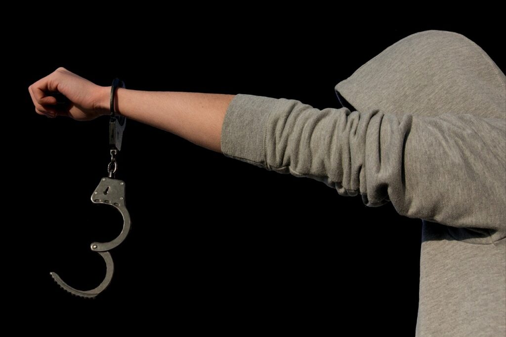 A person in a gray hoodie extends their arm to the side, wearing half of a pair of open handcuffs on their wrist against a black background.
